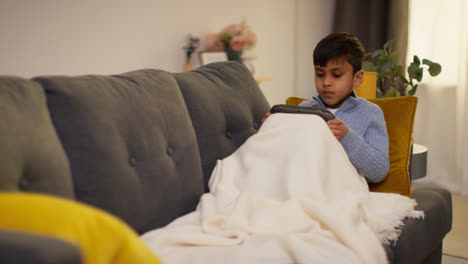 Kleiner-Junge-Sitzt-Zu-Hause-Unter-Der-Decke-Auf-Dem-Sofa-Und-Spielt-Ein-Spiel-Oder-Streamt-Auf-Ein-Tragbares-Spielgerät